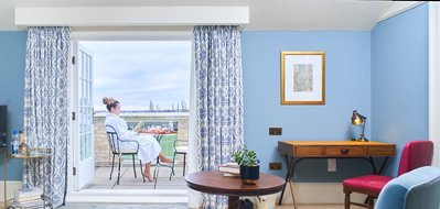 A woman enjoys breakfast on the balcony of a bright and airy hotel room. 