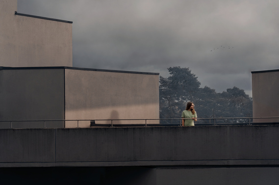 A woman in a pale green dress stands between buildings illuminated by the low sun which casts her shadow on the wall behind her. 