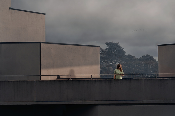 A woman in a pale green dress stands between buildings illuminated by the low sun which casts her shadow on the wall behind her. 