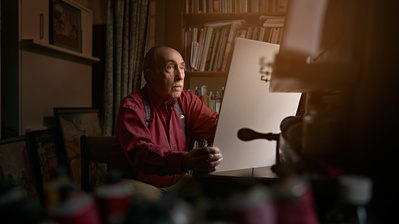 A cinematic portrait of an elder male artist in a vivid red shirt sitting in his painting studio, lit up by warm sun light coming through the window in front of him. 