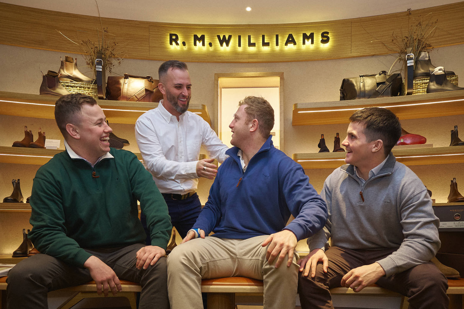 Three people sat down in a mens shoe shop being helped by the shop keeper. 