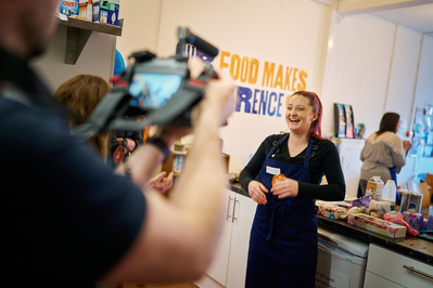 A portrait of a woman laughing, wearing an apron whilst a film crew interviews her. 