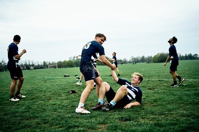 A group of boys are playing rugby. One of the boys has been tackled to the ground and another boy is helping him up. 