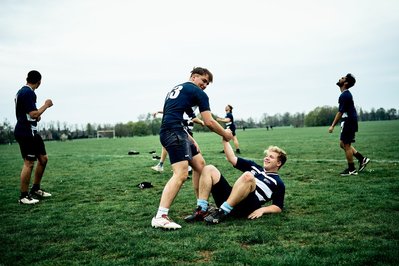 A group of boys are playing rugby. One of the boys has been tackled to the ground and another boy is helping him up. 
