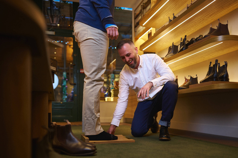 A man is being helped by a shop keeper in a shoe shop. 