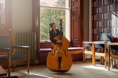 A young girl in a library playing the cello, backlit by the sun. 