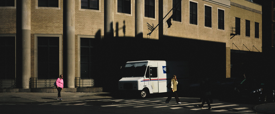 People walking the street as a parcel delivery van comes out of the shadows. 