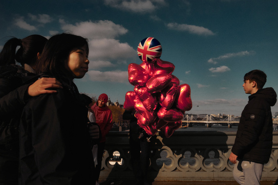 A man is selling vibrant red balloons on a London bridge as pedestrians walk by. 
