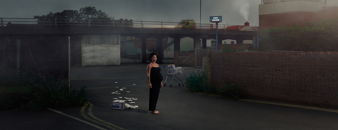 a cinematic and dramatic advertising photograph of a pregnant woman standing alone with a shopping trolley behind her at twilight surrounded by buildings in an urban setting. 