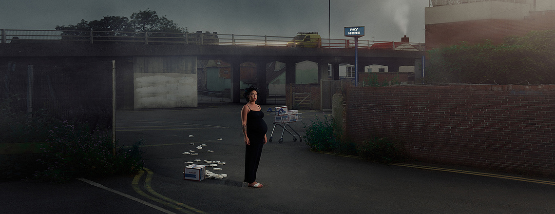 a cinematic and dramatic advertising photograph of a pregnant woman standing alone with a shopping trolley behind her at twilight surrounded by buildings in an urban setting. 