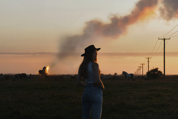 A cow girl stands in a field with horses as a building in the distance is on fire. 