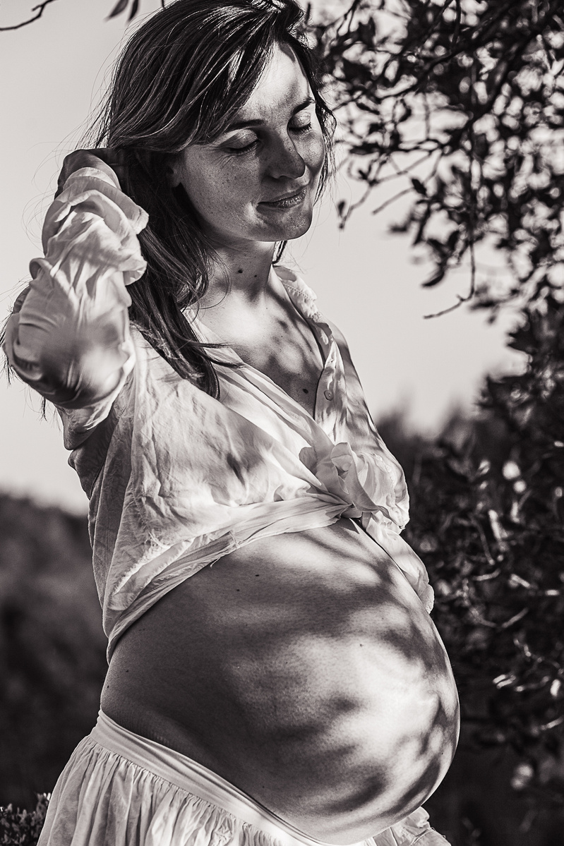 Mujer embarazada se toca el pelo y deja entrever su barriga con sombras y reflejos de la naturaleza en playita de l'estany.