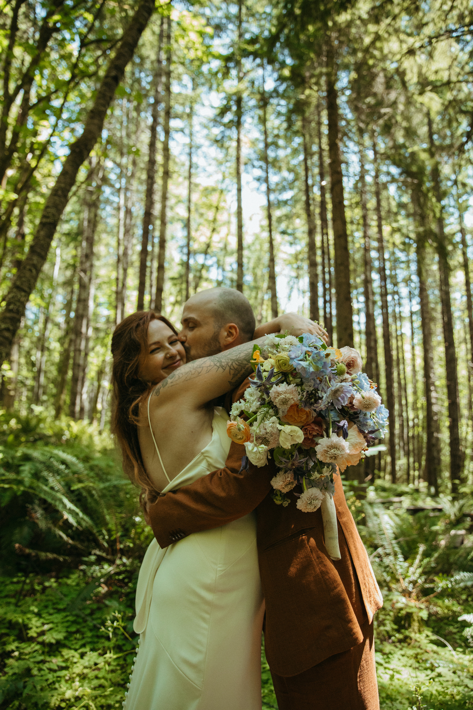 An indie couple snuggles underneath the PNW forest on their wedding day with a big beautiful bouquet near Mt. Rainier Washington.