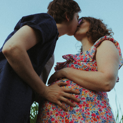 A pregnant queer couple taking their maternity photos underneath a bright blue sky in the Summer in Minnesota.