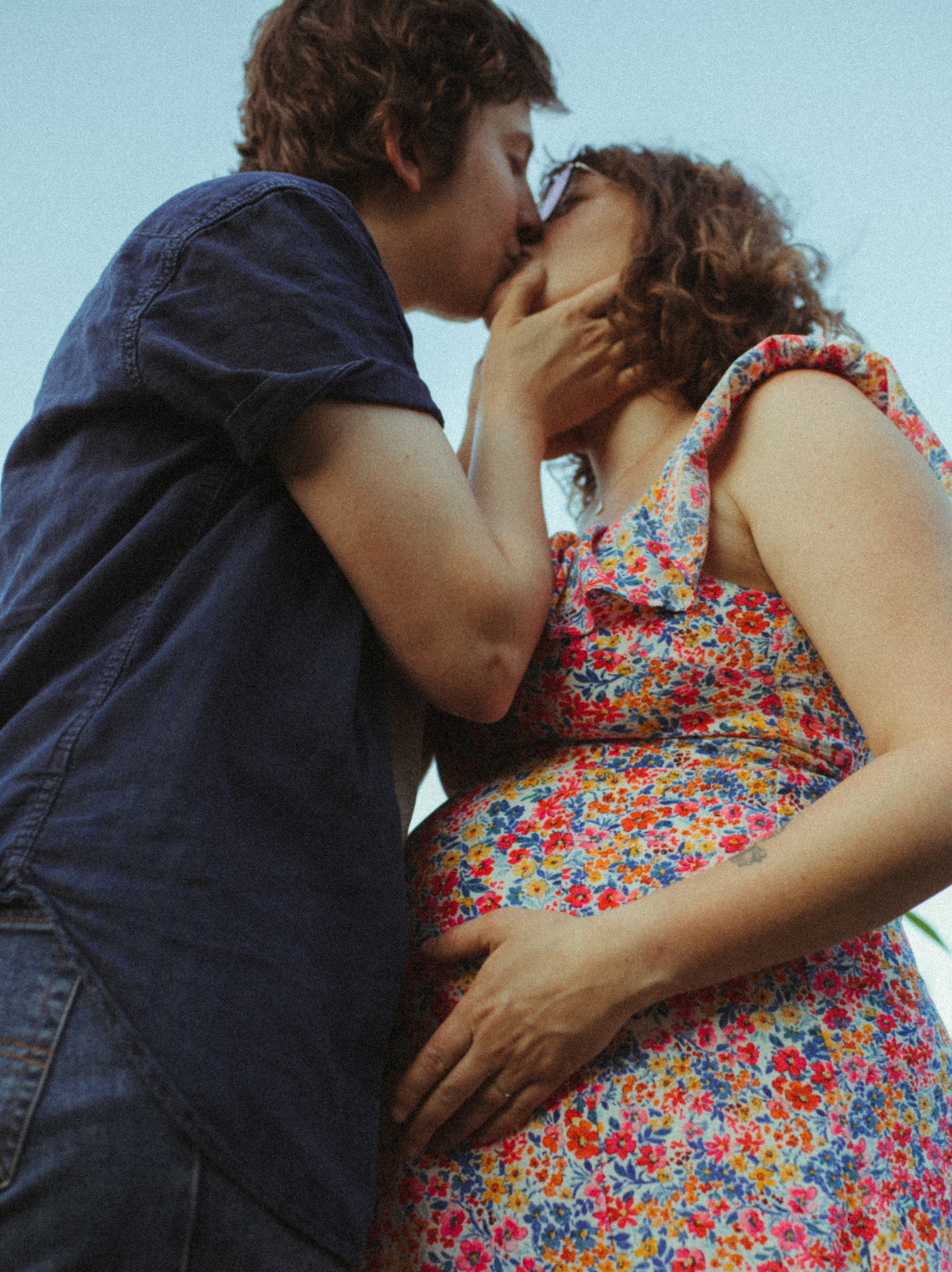 A queer couple kisses and touches belly bump during their maternity shoot underneath a bright blue sky.