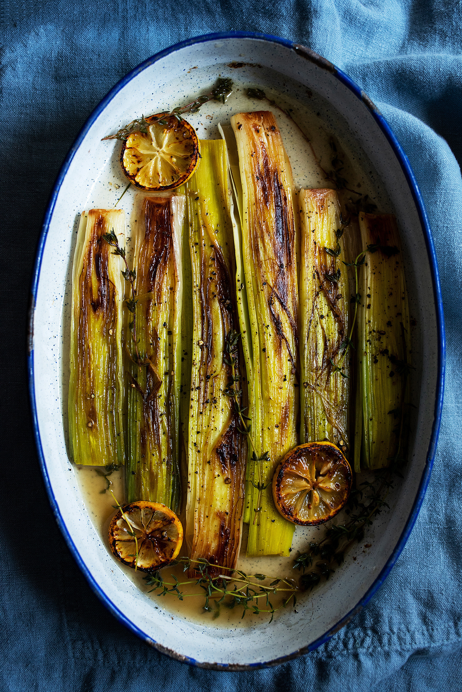 Braised leeks in an oval roasting tin shot from overhead on a blue linen background.