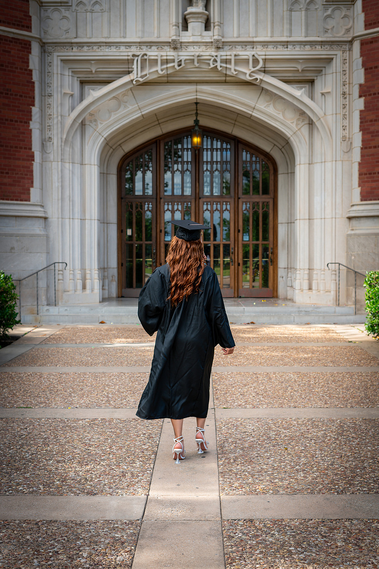 College graduate wearing cap and gown and walking toward University of Oklahoma library.