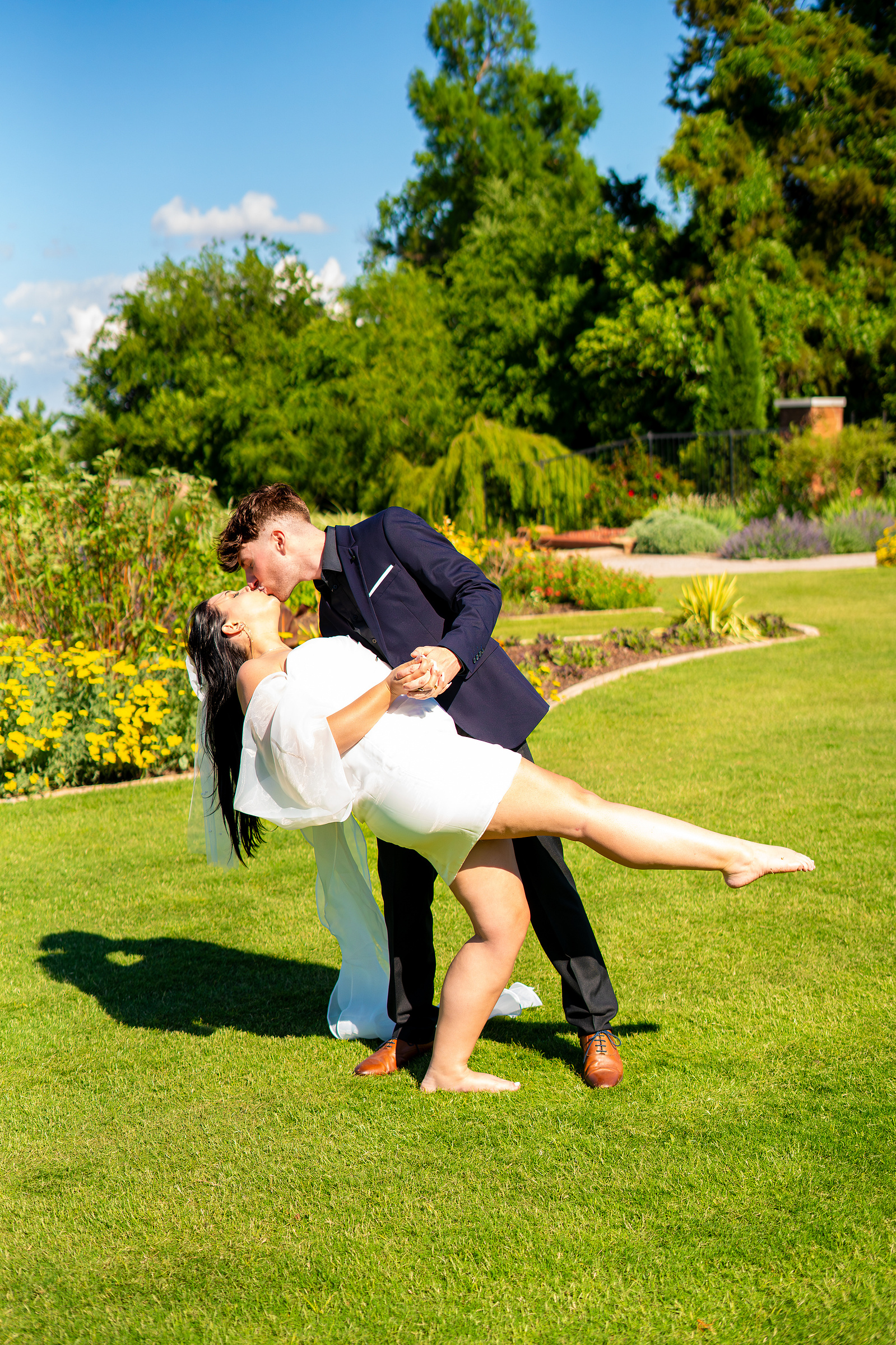 A romantic outdoor wedding pose as the groom, in a navy suit, gracefully dips his barefoot bride on a lush green lawn. The vibrant garden setting adds to the dreamy, sunlit atmosphere.