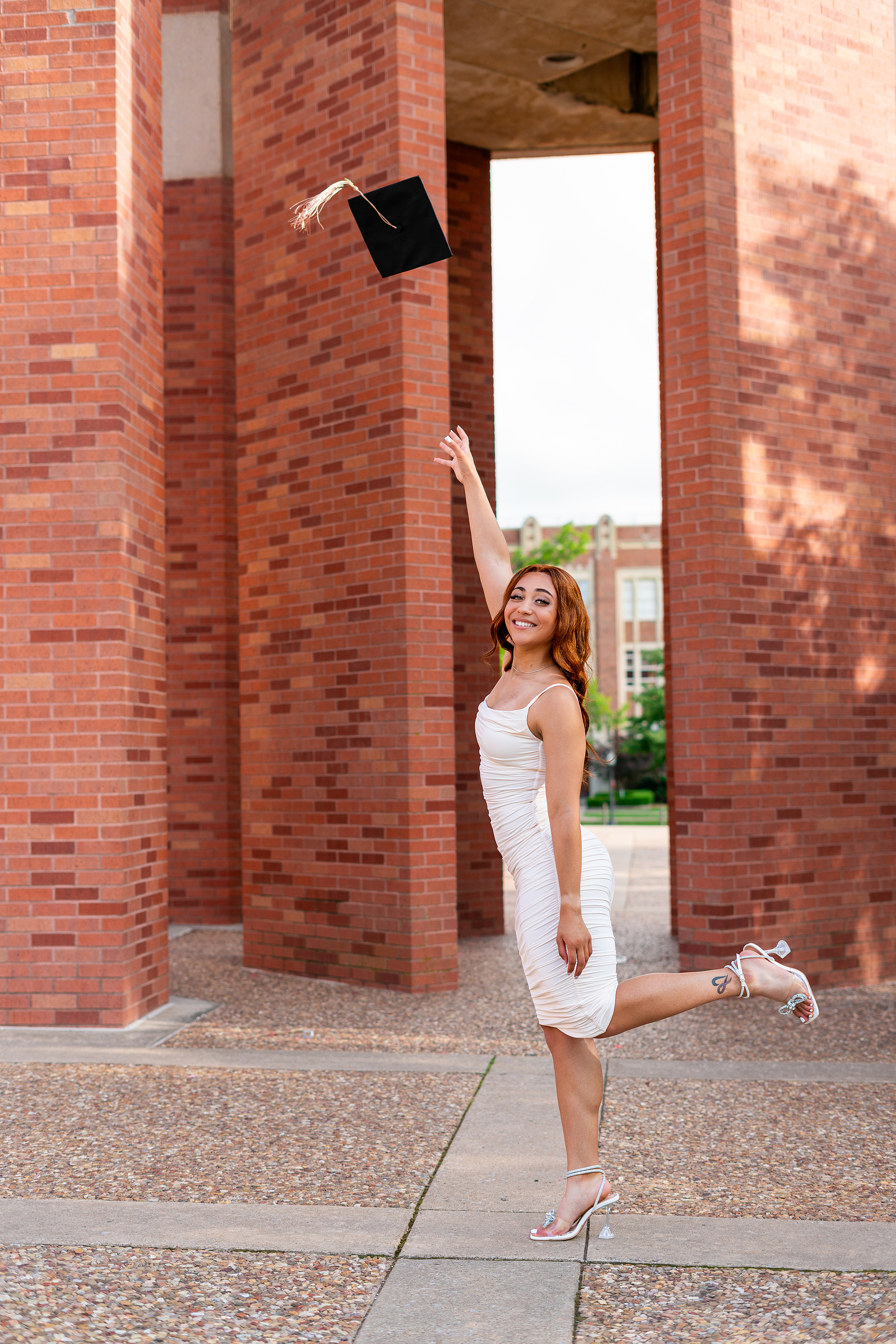 A joyful graduate in a white dress throws her cap in the air, celebrating her achievement in front of a striking red brick structure.