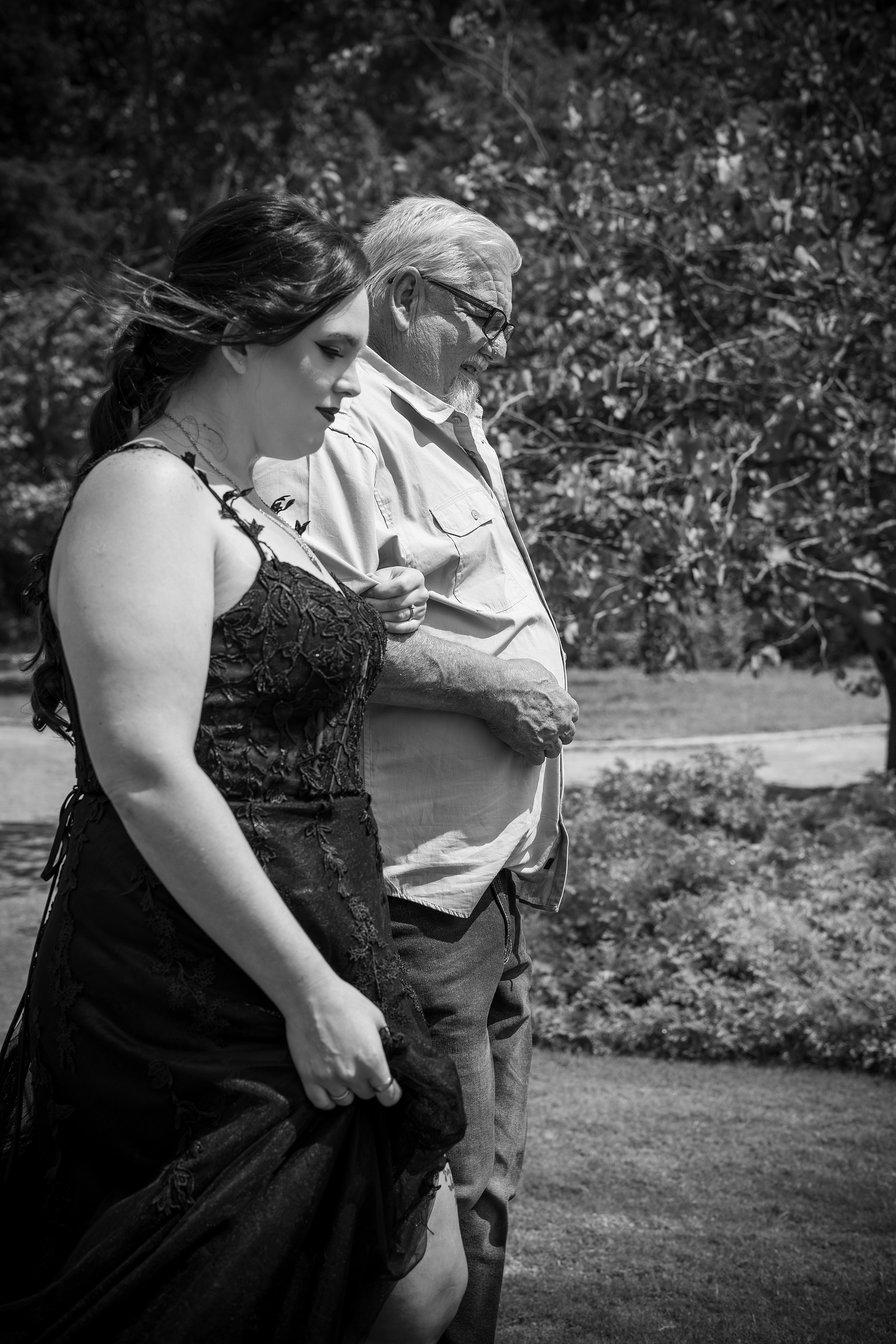 A bride in a black lace wedding dress walks alongside an older man in an emotional black-and-white moment wedding moment.