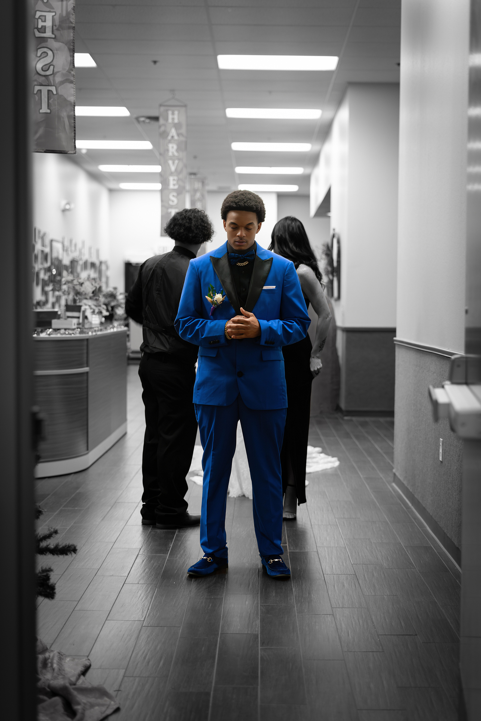 A young man in a sharp blue suit stands confidently in a modern hallway, the selective color emphasizing his striking outfit. 