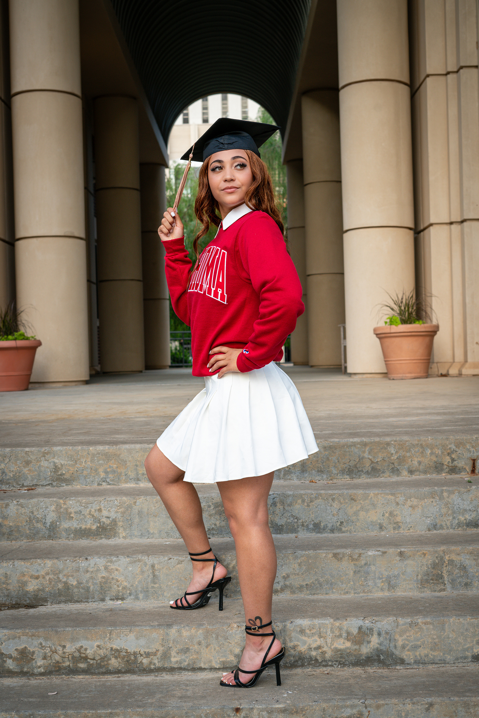 A confident graduate poses on stone steps at the University of Oklahoma, wearing a red sweater, white skirt, and a black graduation cap, symbolizing achievement and celebration.