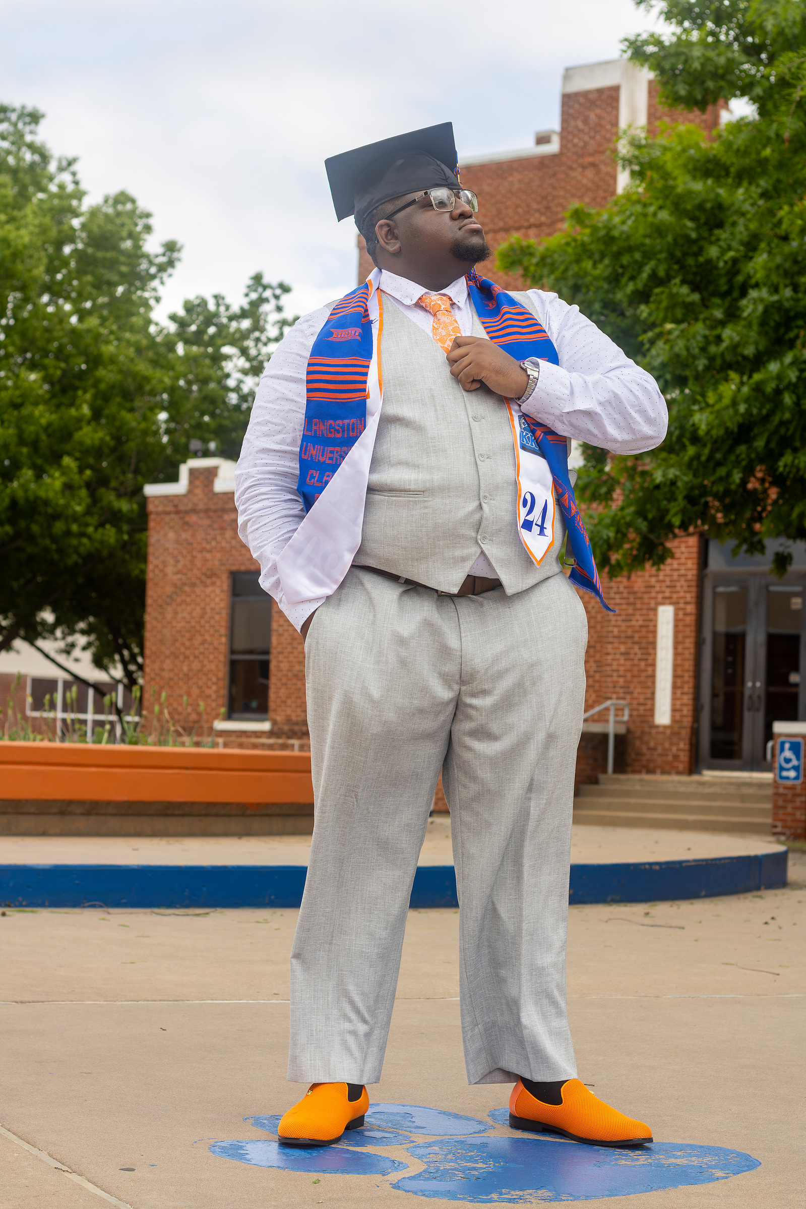 A proud graduate in a light gray suit, orange accents, and a traditional stole gazes into the distance, standing on a university campus.