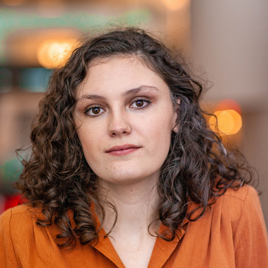 Woman wearing orange shirt with brown hair and eyes in front of abstracted background.