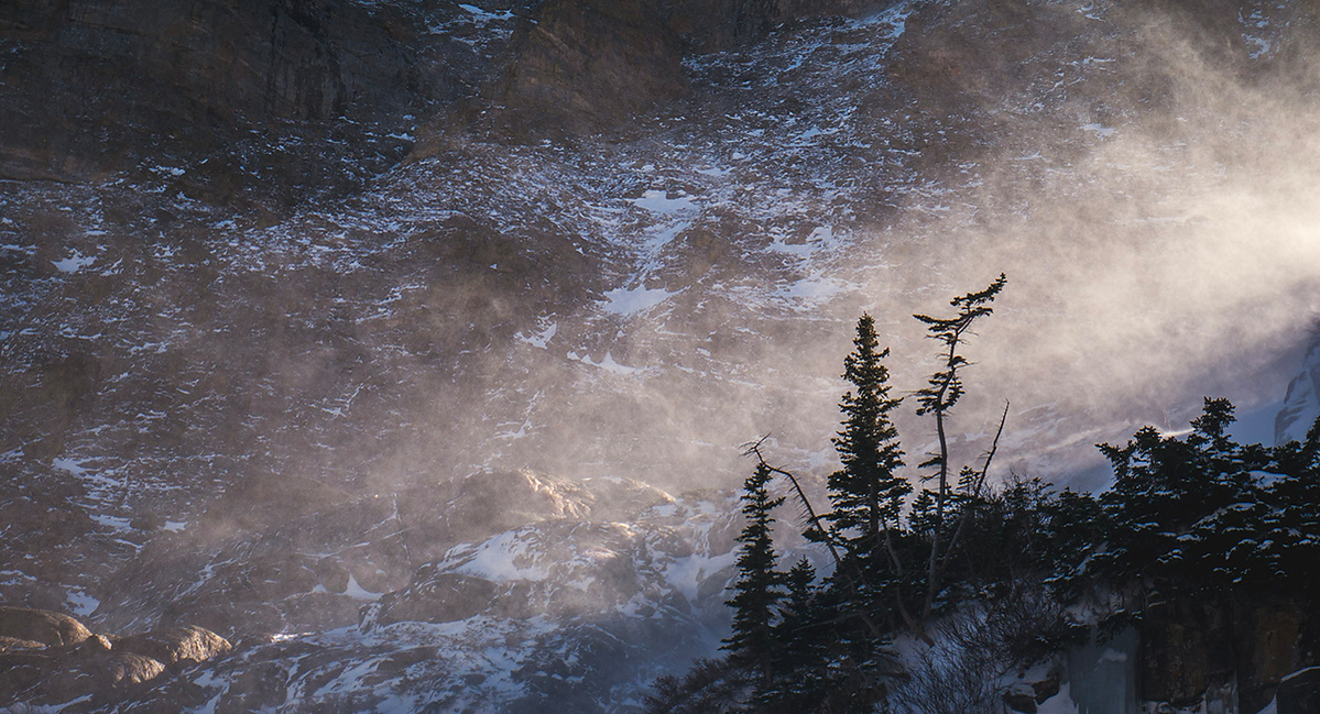 Landscape photograph of blowing snow lit by the sun with tree silhouettes in front and mountainside in the background