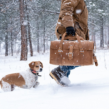 Man in large brown coast carrying a large leather bag though the snow, while it is snowing, with forest in the background. A hunting dog follows and looks at the camera. This speaks directly to the brand's target market.