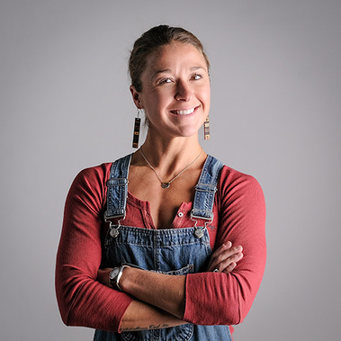 Studio portrait of woman in overalls and red shirt, with arms folded, shot in studio with semi-hard light. This is a great headshot, and could be used for social media, or a personal or professional web site.
