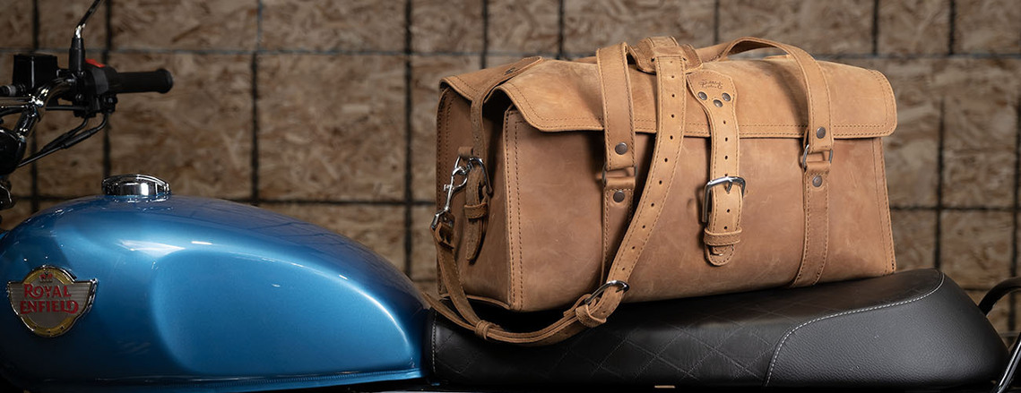 Tan leather bag on motorcycle seat in maintenance shop. Product shows a possible use, and speaks to the lifestyle of the company's target market.