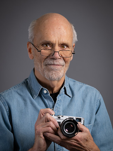 Studio portrait of older gentleman, in blue denim shirt, holding camera. Portrait uses a studio background. His glasses pose only a minor issue which can be handled easily. This is perfect for social media, or other professional use.