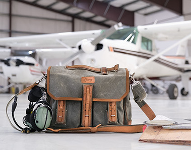 Styled product shot of green canvas flight bag, with pilot's accessories, in aircraft hangar, and aircraft. Shows intended use, and appeals to the manufacturer's target market.