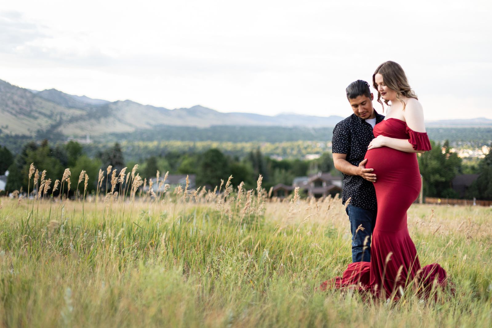 A pregnant woman in an elegant red gown with her husband holding her stomach, overlooking the Boulder, Colorado foothills and Colorado plains during a Maternity Photoshoot. 