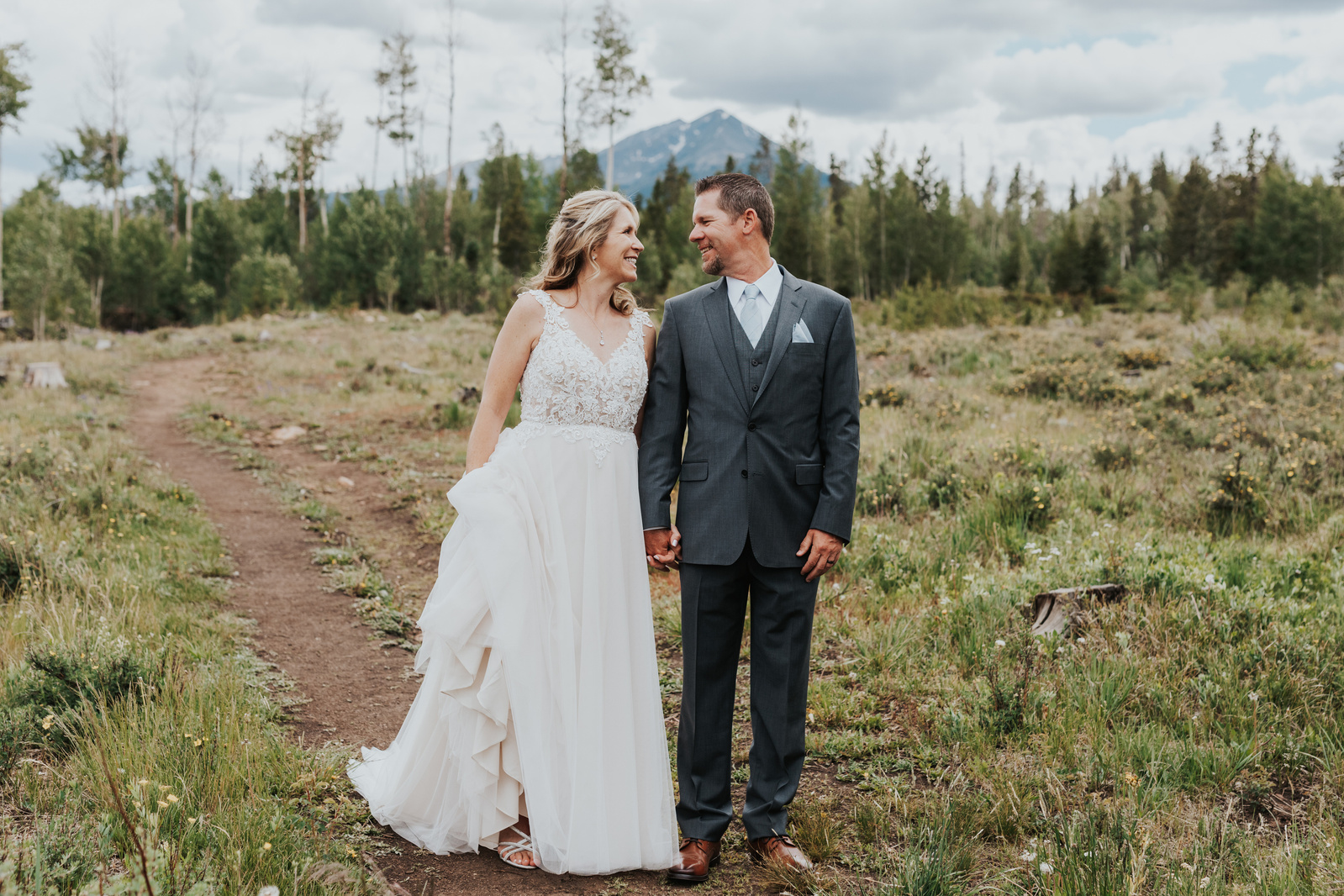 Bride and Groom stand on a trail in Silverthorne, Colorado with a mountain backdrop on their wedding day at the Silverthorne Pavillion. Photograph taken by Denver Wedding Photographer, Simone Schiess. 
