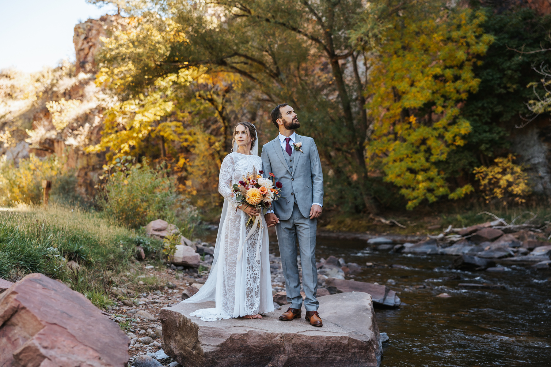 Colorado Wedding Photography featuring Caity & Mike at the River Bend at Lyon's Farmette is Lyon's Colorado, near Boulder Colorado. Bride & Groom are standing on a rock in a river in the fall with orange and yellow trees behind them changing color. 