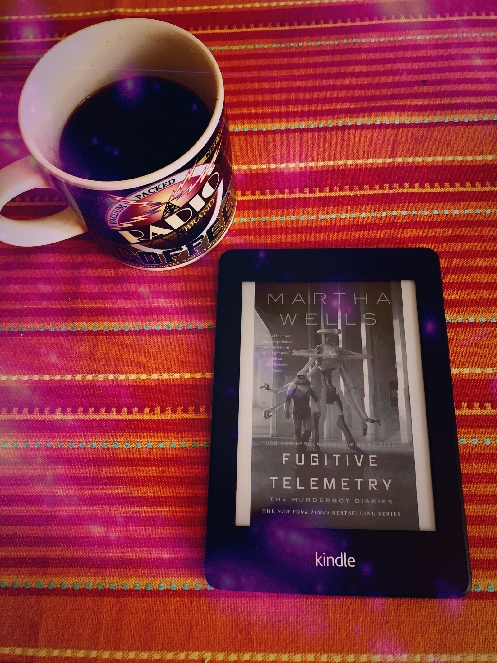 A kindle displaying the cover for Fugitive Telemetry, by Martha Wells, lies on a red, orange and yellow striped tablecloth next to a fresh cup of Moka pot coffee. They're both mine, you can't have them.