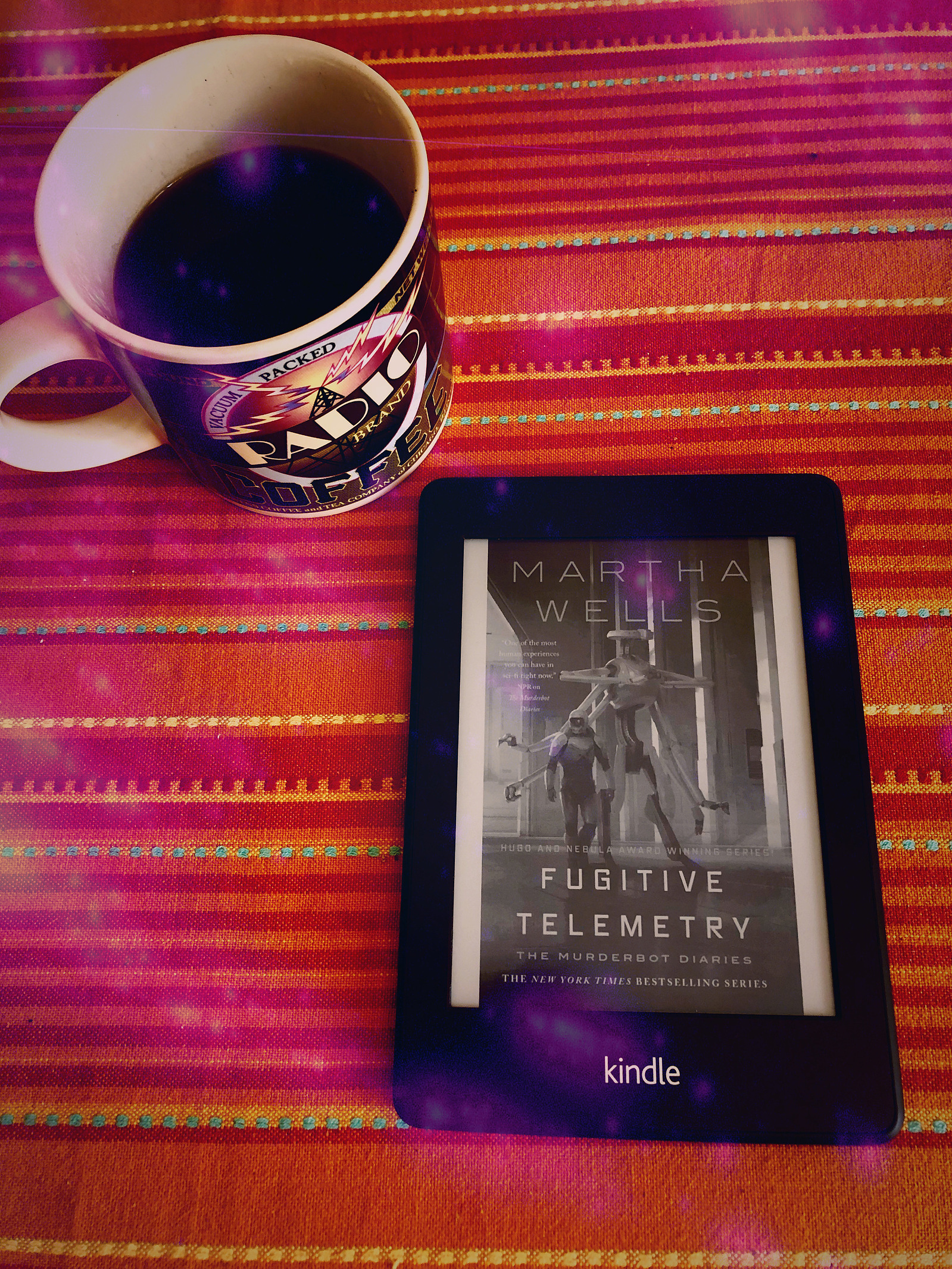 A kindle displaying the cover for Fugitive Telemetry, by Martha Wells, lies on a red, orange and yellow striped tablecloth next to a fresh cup of Moka pot coffee. They're both mine, you can't have them.