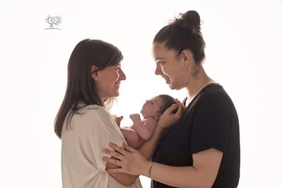 Pareja de mamas posando con su bebe en brazos, situadas de perfil a la camara y mirandose. Fotografia profesional