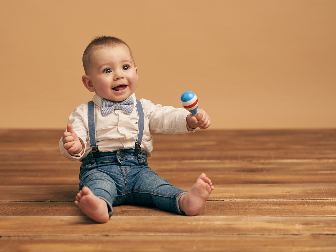 Bebe risueño vestido con camisa blanca tirantes y vaqueros. Sujetando una maraca. Fotografía profesional en Gijón