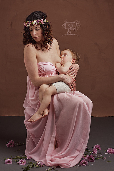 Mama con diadema y vestido de vuelo dando el pecho a su hijo en un escenario con flores