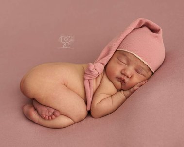Recien nacido con gorrito dormilon durmiendo sobre tela color beige y la mejilla apoyada en las manos