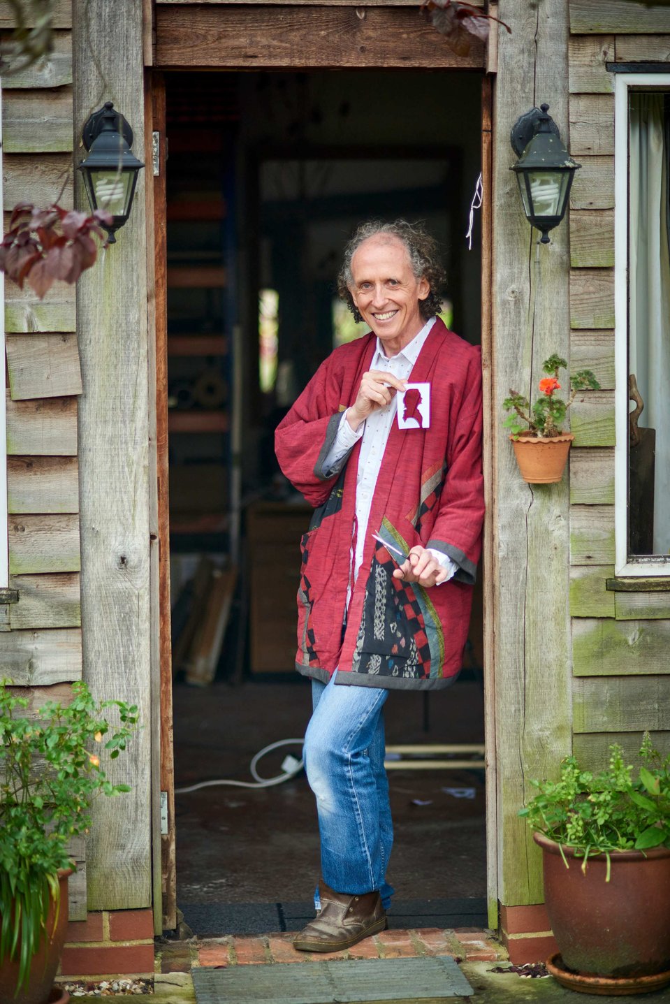 Man standing in doorway in red coat with stencil and scissors in hand.
