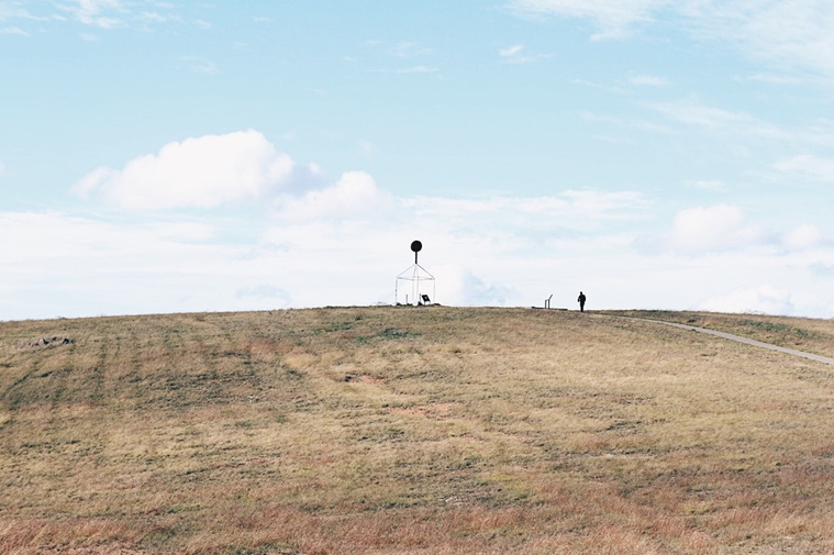 A landscape shot of a grassy hill with a navigation structure on the knoll. A solitary figure walks up the path to the top. It is a bright and cloudy day. 