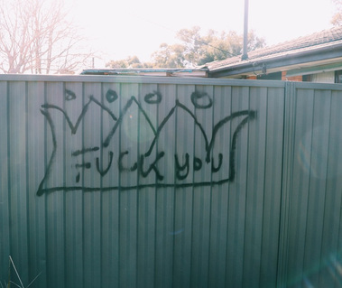 Graffiti on a corrugated walk. Black outline of a crown with 'fuck you' written inside it. 
