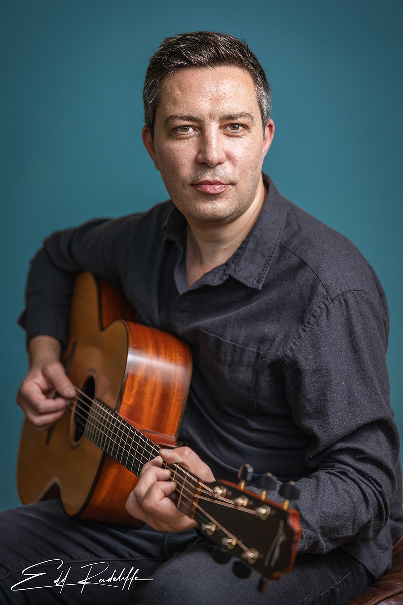 Portrait of a man in a dark shirt playing an acoustic guitar against a teal background. Ideal for musician profiles, promotional materials, or artistic branding.