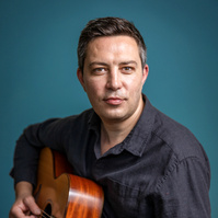 Portrait of a man in a dark shirt playing an acoustic guitar against a teal background. Ideal for musician profiles, promotional materials, or artistic branding.