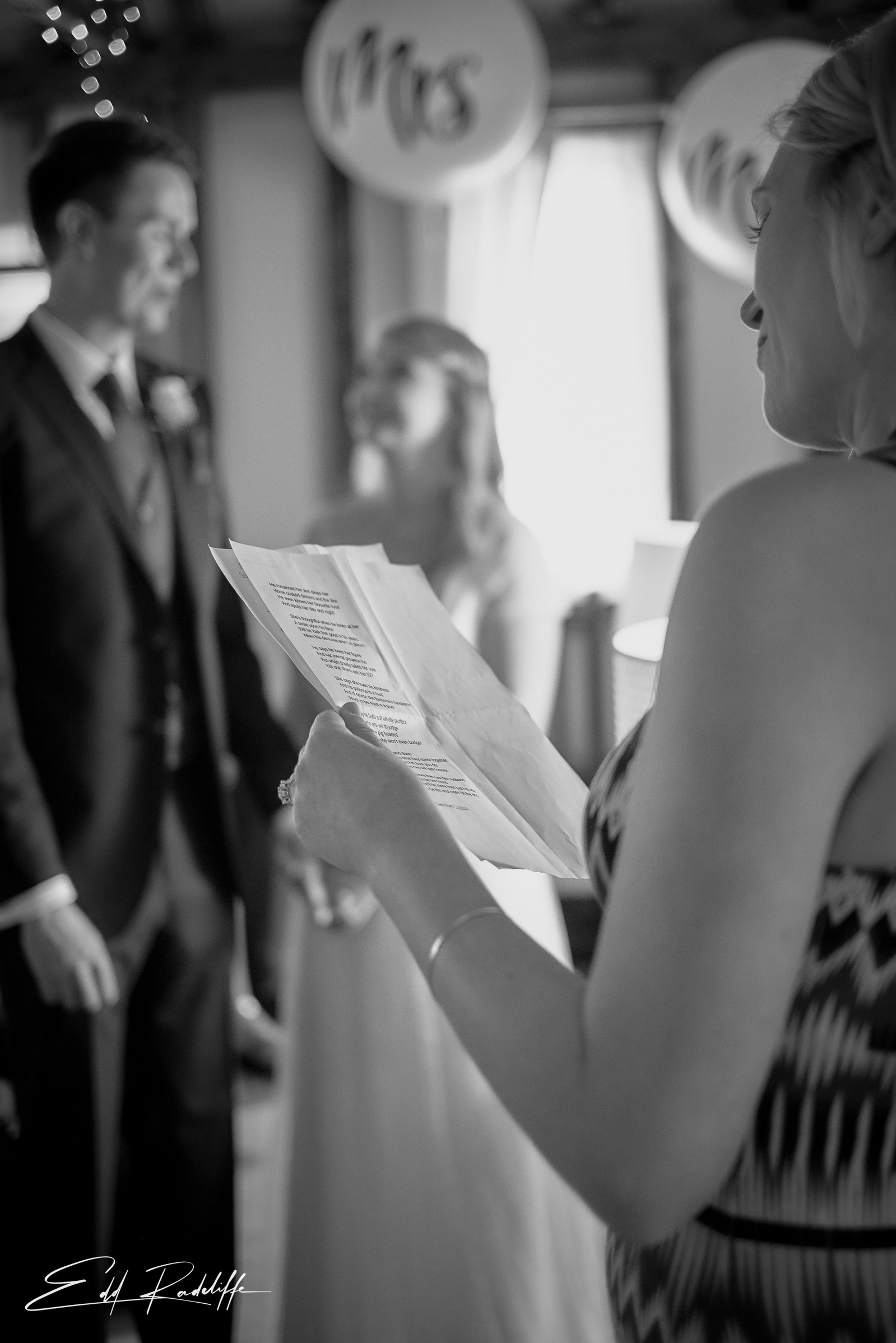 Bride and Groom smile at each other while guest does reading during wedding ceremony at the Reid Rooms, Chelmsford for Edd Radcliffe Photography - documentary style wedding photography