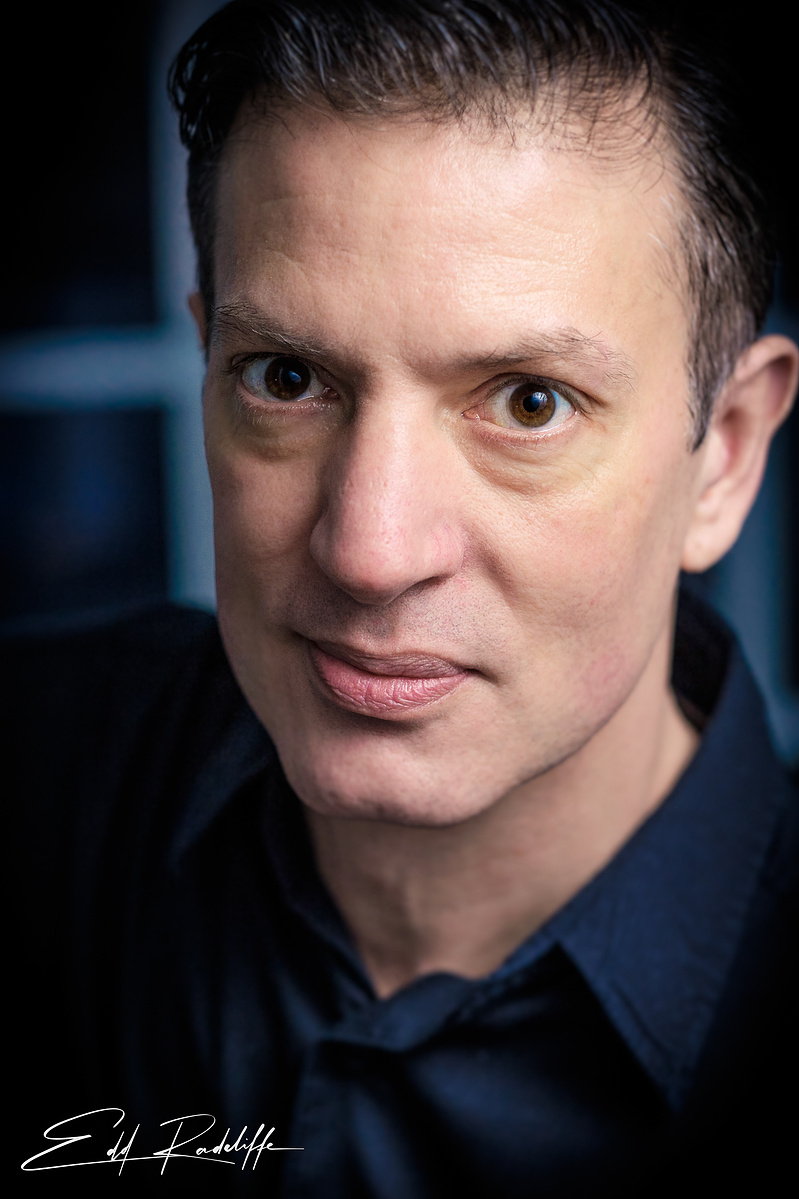Close-up professional headshot of a man in a dark shirt with an intense expression, set against a softly lit, dark background. Perfect for actor profiles or creative branding.